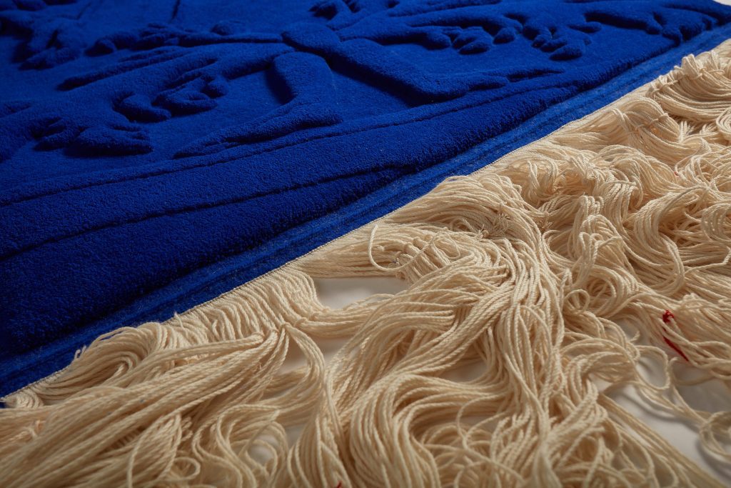 Tapisserie/tapis fait à la main selon la technique de la Savonnerie avec au premier plan les chaînes de laine écrues, au second plan la lisière et le velours travaillé en relief de la pièce Métamorphose IV. L'écru des chaînes tranche avec le bleu du tissage.